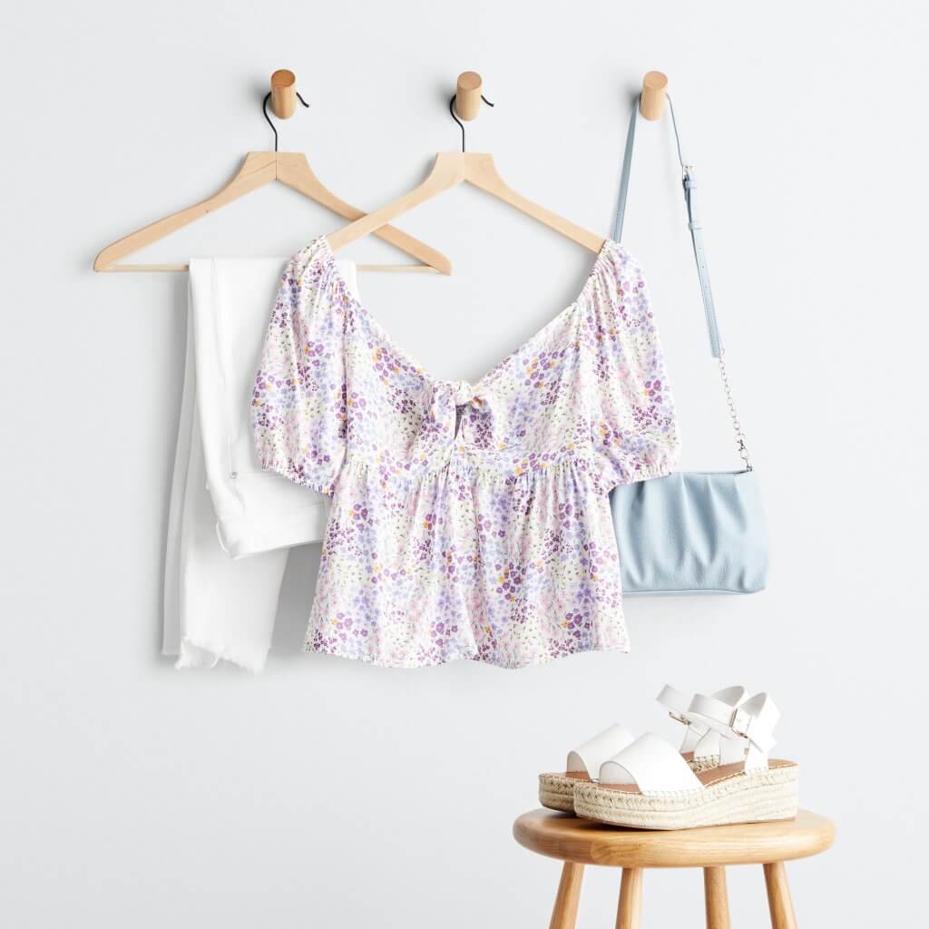 Stitch Fix women’s luxury fabric purple floral print blouse, white jeans and blue crossbody bag on hangers next to white espadrille platform shoes on stool.