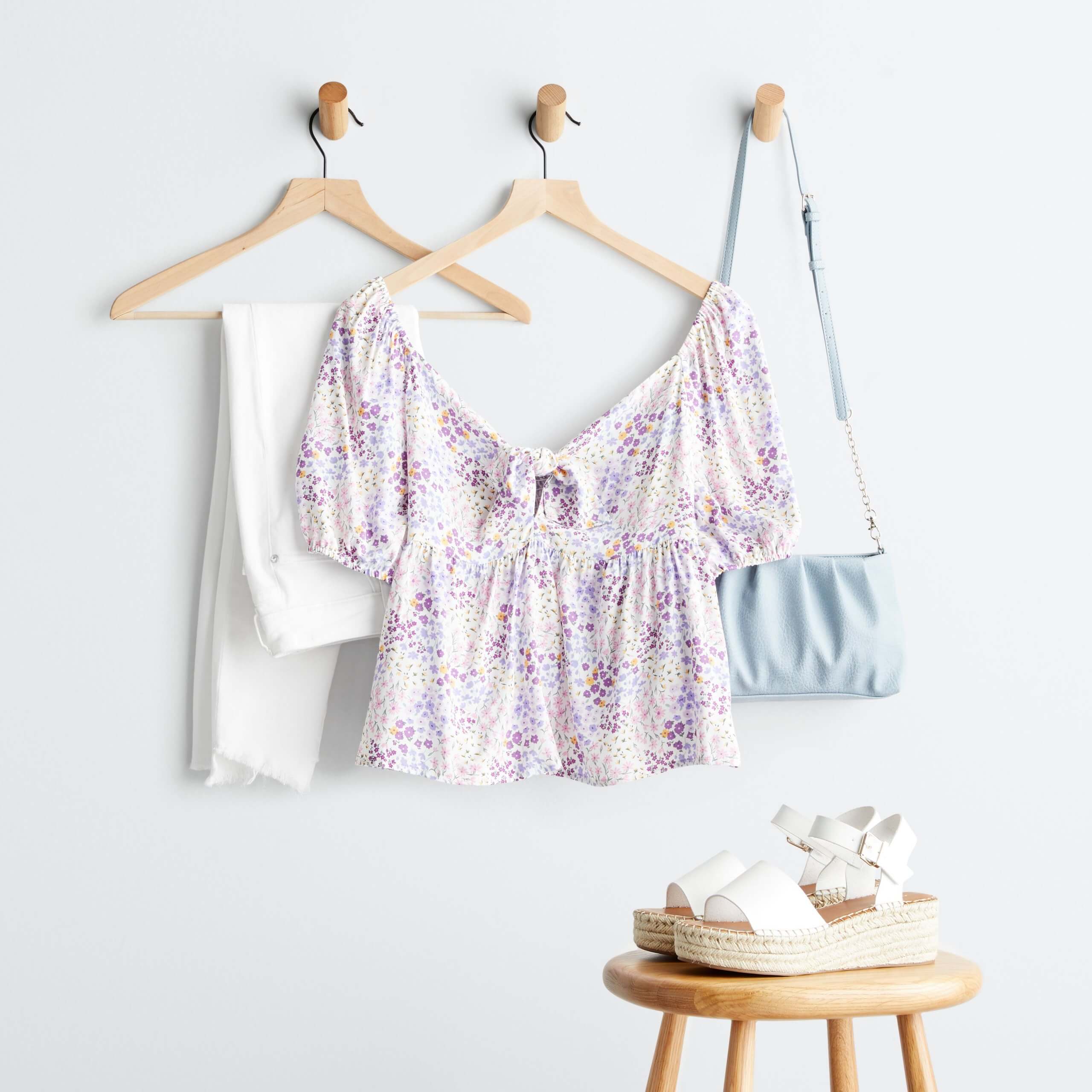 Stitch Fix women’s luxury fabric purple floral print blouse, white jeans and blue crossbody bag on hangers next to white espadrille platform shoes on stool.