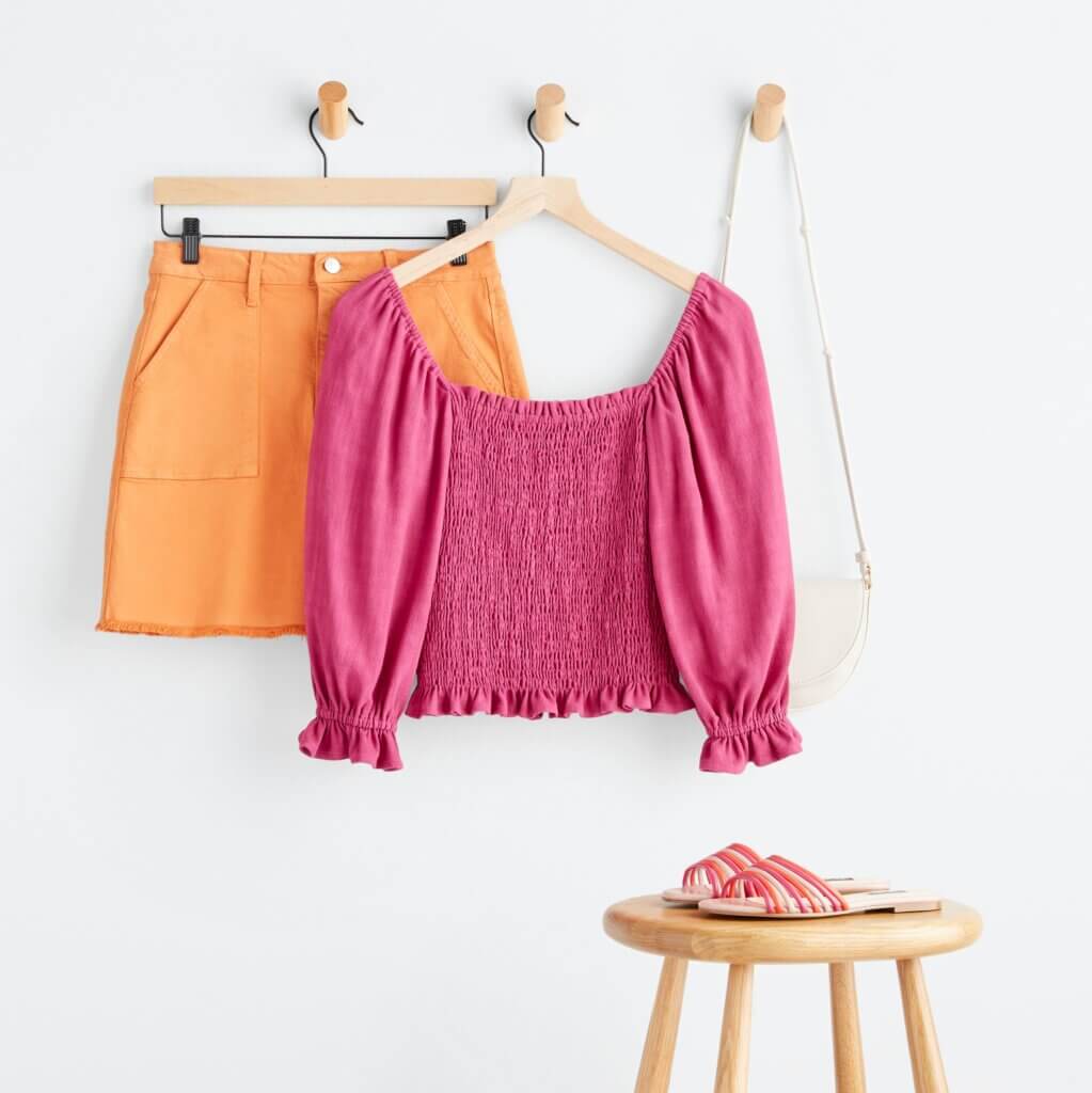 Stitch Fix Women’s outfit featuring orange denim skirt, pink off-the-shoulder top, white purse hanging on hooks next to pink sandals on a stool.