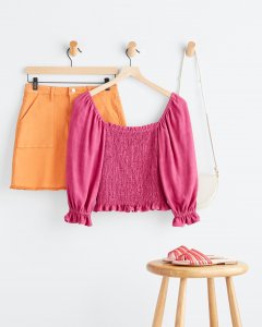 Stitch Fix women’s orange mini skirt, pink puff sleeve top and a white purse hanging behind a stool with pink sandals on top.