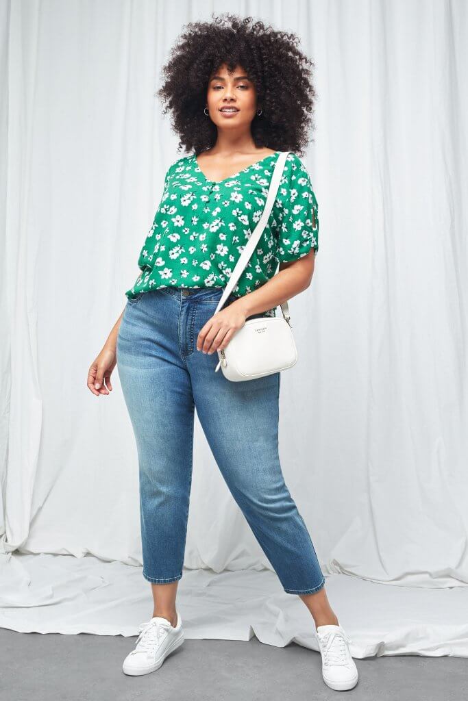 Stitch Fix model wearing plus-size women’s green floral blouse, blue denim capris, white sneakers and white crossbody bag.