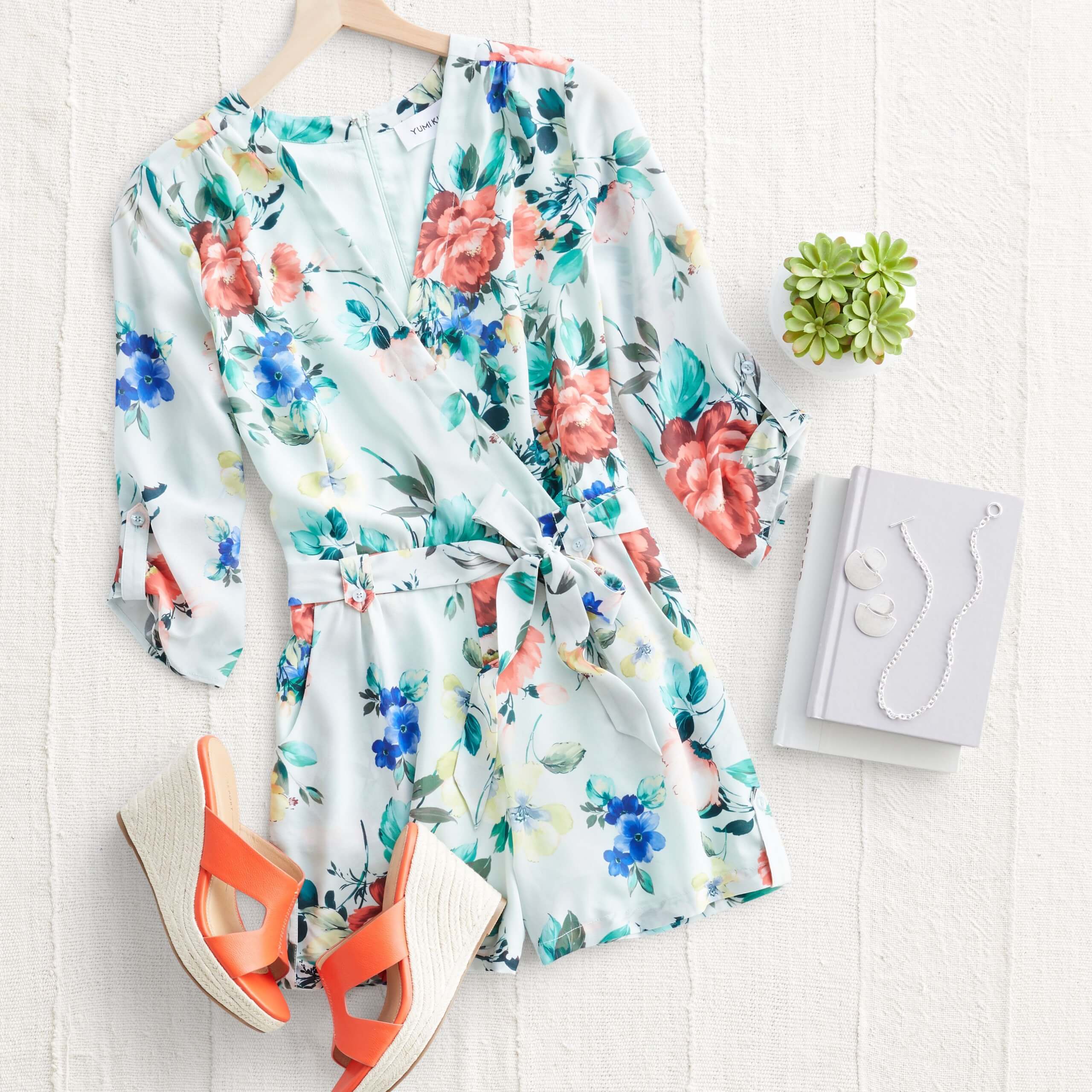 Stitch Fix Women's outfit laydown featuring white floral romper, coral wedges and silver drop earrings and necklace on white book.