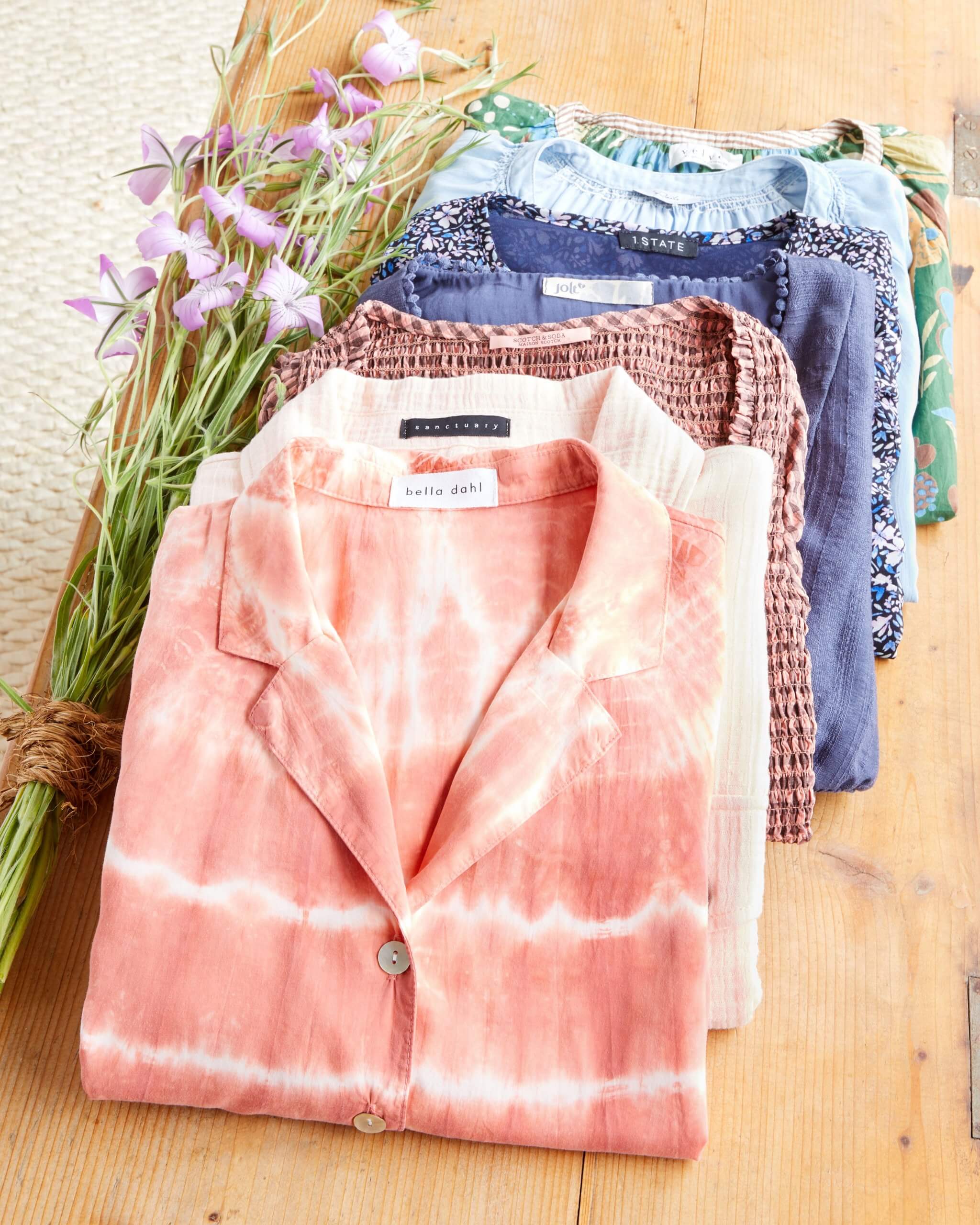 Stitch Fix Women’s outfit laydown featuring folded shirts in a pile in pink, white, purple, blue and green tones on a wooden table. 