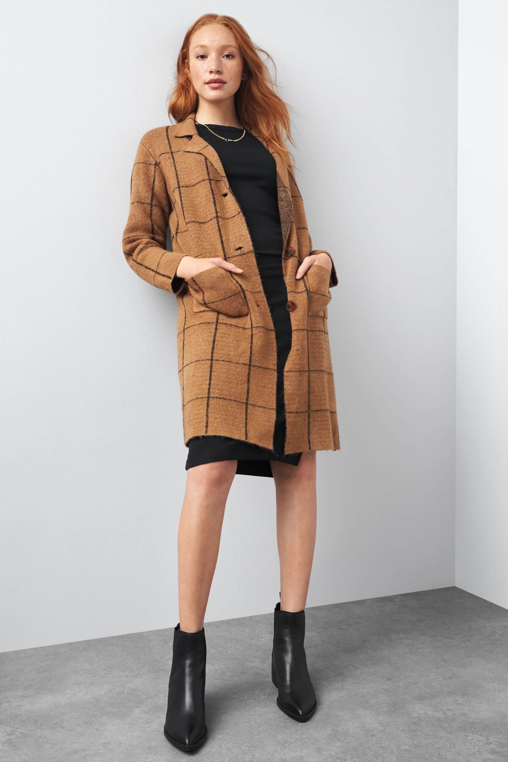 Stitch Fix women’s model in a dark academia aesthetic outfit featuring a black dress, tan windowpane-patterned cardigan and black booties.