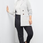 Stitch Fix women’s model wearing black trousers, white turtleneck knit top, grey double-pocket coatigan, and black loafers leaned against a white wall.