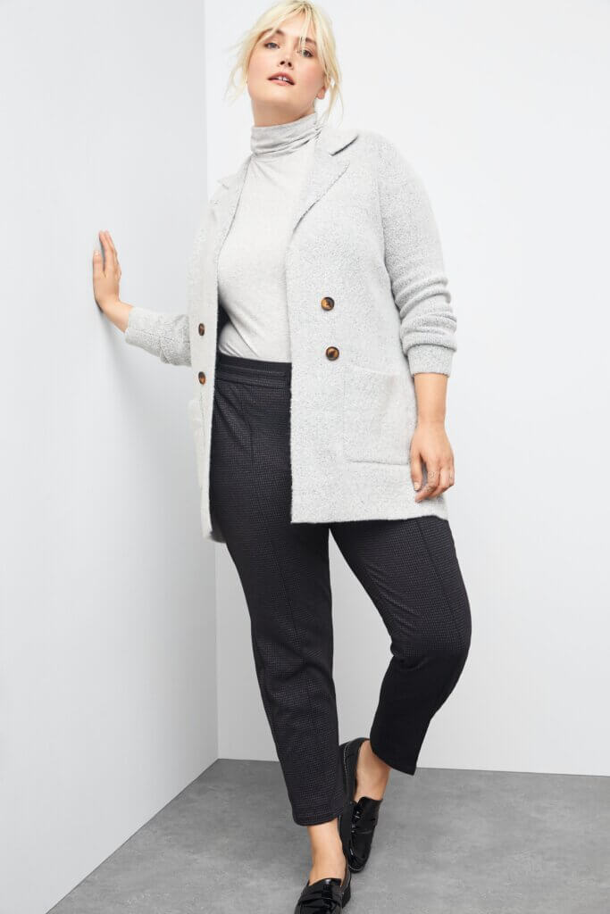 Stitch Fix women’s model wearing black trousers, white turtleneck knit top, grey double-pocket coatigan, and black loafers leaned against a white wall.
