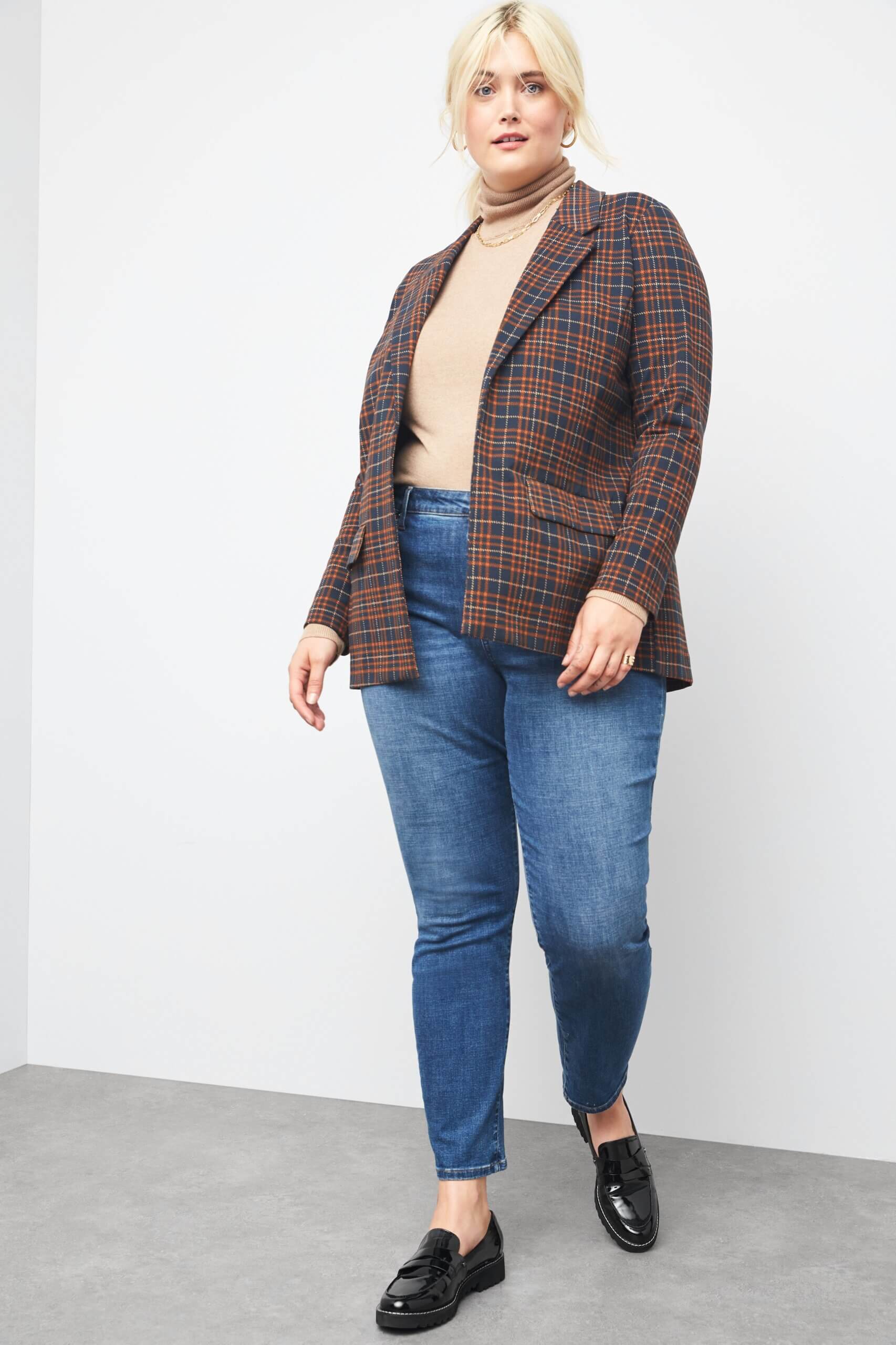 Stitch Fix women’s model wearing plus size business casual outfit with plaid blazer, beige turtleneck, blue skinny jeans, black loafers and gold accessories.