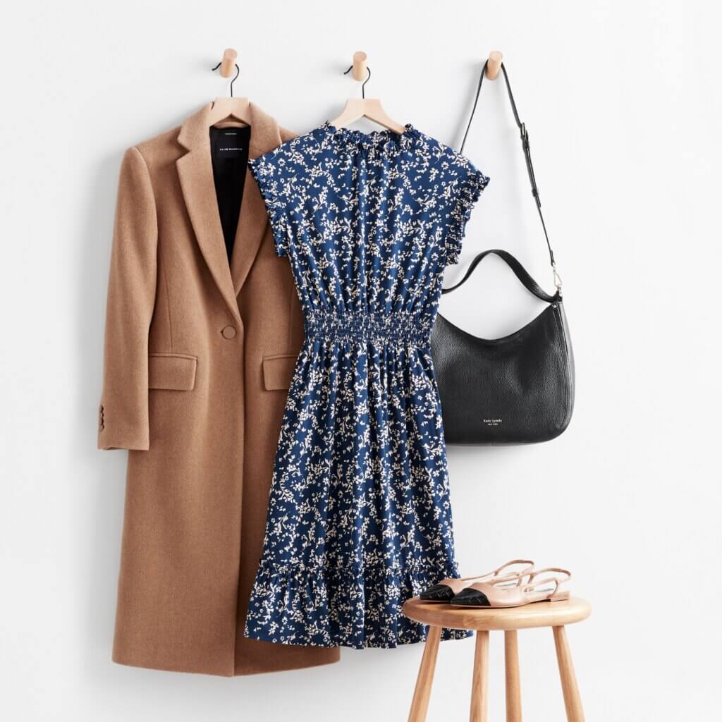 Stitch Fix women’s winter dress outfit with camel overcoat, navy printed dress and black purse on hooks next to color-blocked flats on stool.