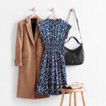 Stitch Fix women’s winter dress outfit with camel overcoat, navy printed dress and black purse on hooks next to color-blocked flats on stool.