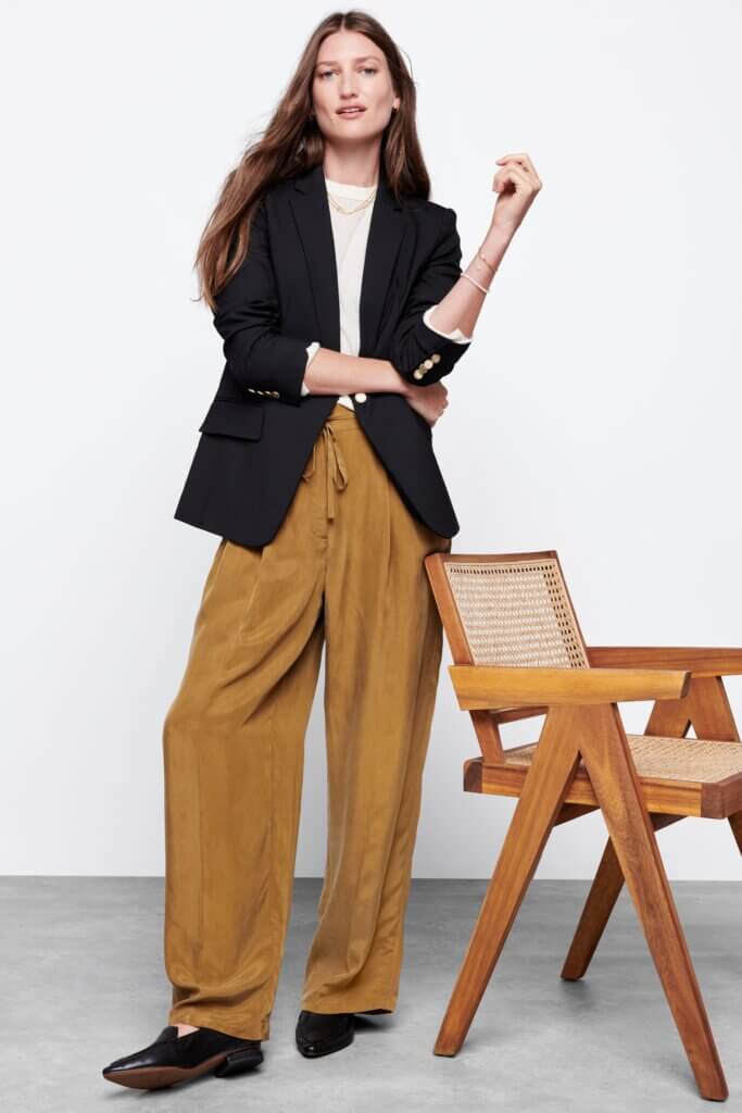 Stitch Fix model in normcore aesthetic outfit for women featuring white crew neck sweater, black blazer, brown wide-leg trousers and black loafers.