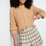 Stitch Fix women’s model in a dark academia outfit featuring a tan cable knit sweater and beige and grey plaid skirt.