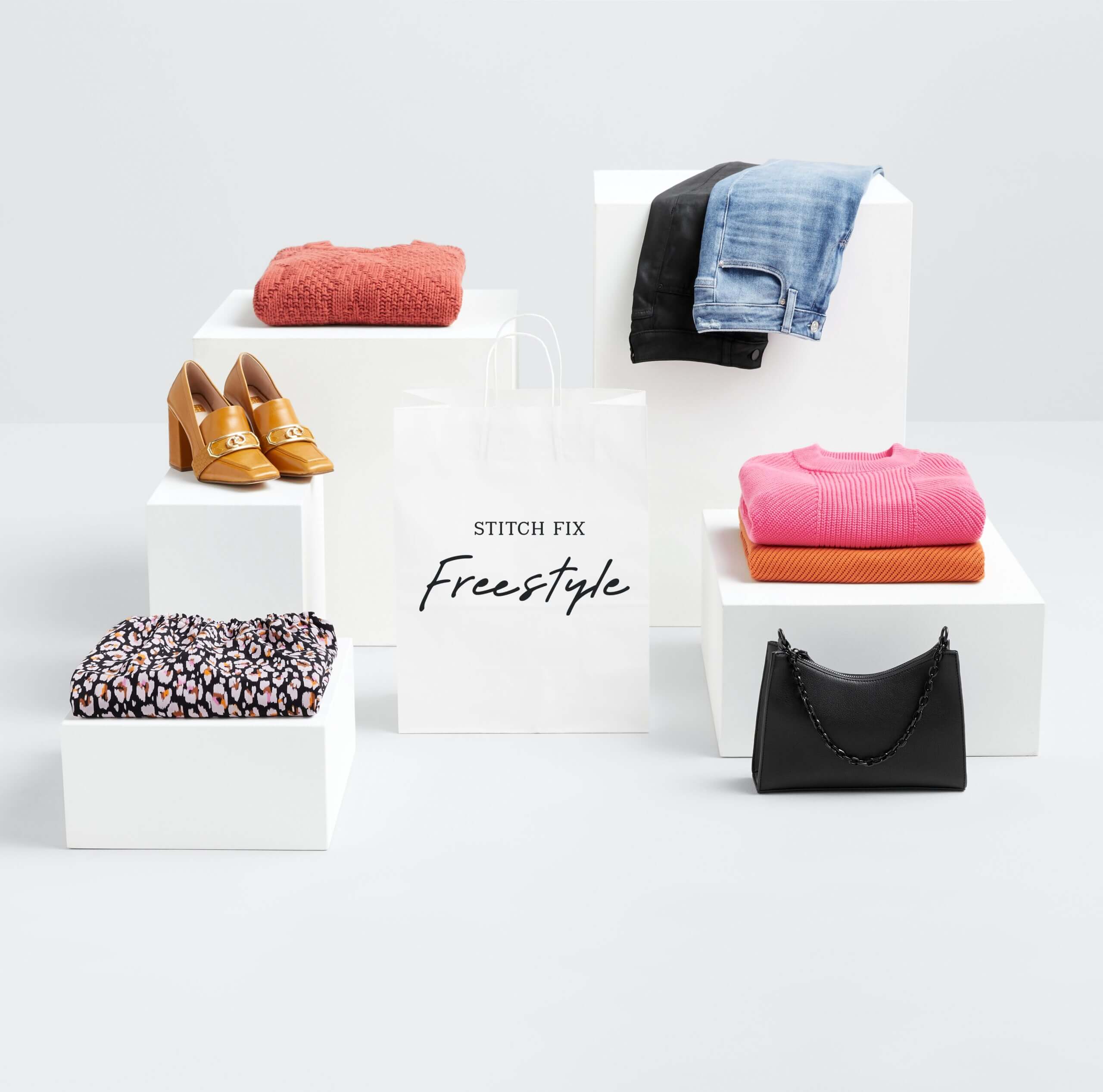 Stitch Fix 2022 fashion trends for women display featuring blue jeans, black pants, bright folded sweaters, floral folded blouse, heeled loafers and black bag with shoulder strap.