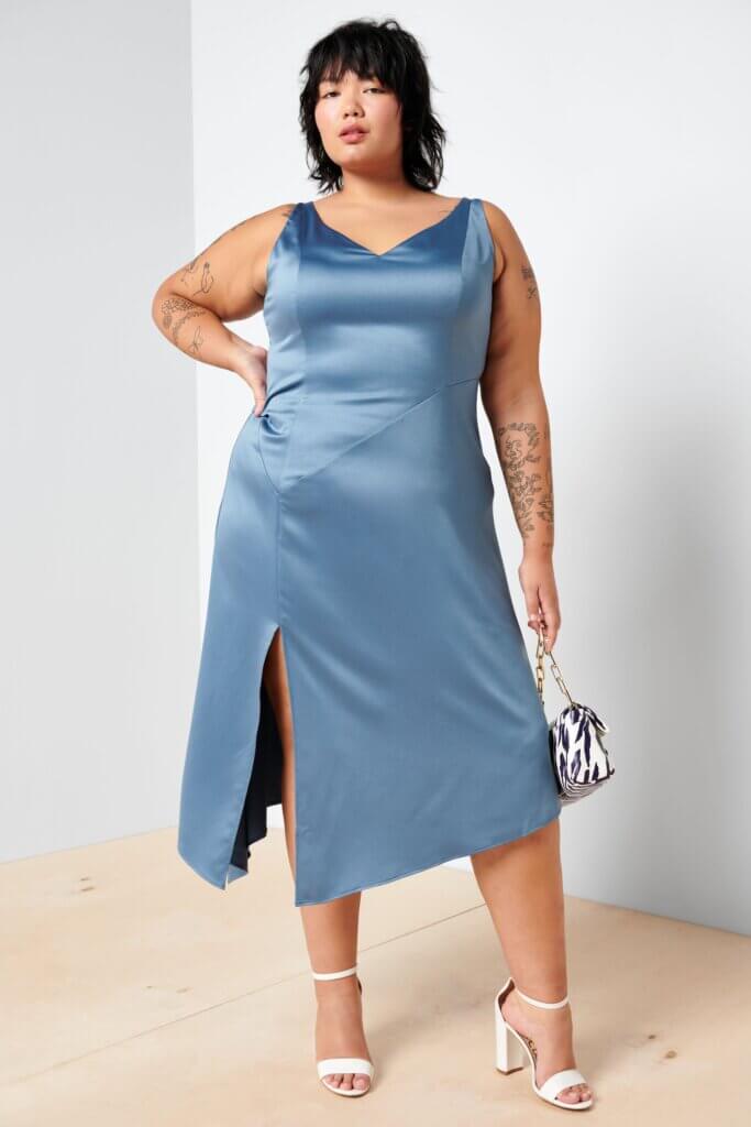 Stitch Fix model wearing plus size women’s semi-formal wedding attire outfit with blue satin dress, printed handbag and white heels.