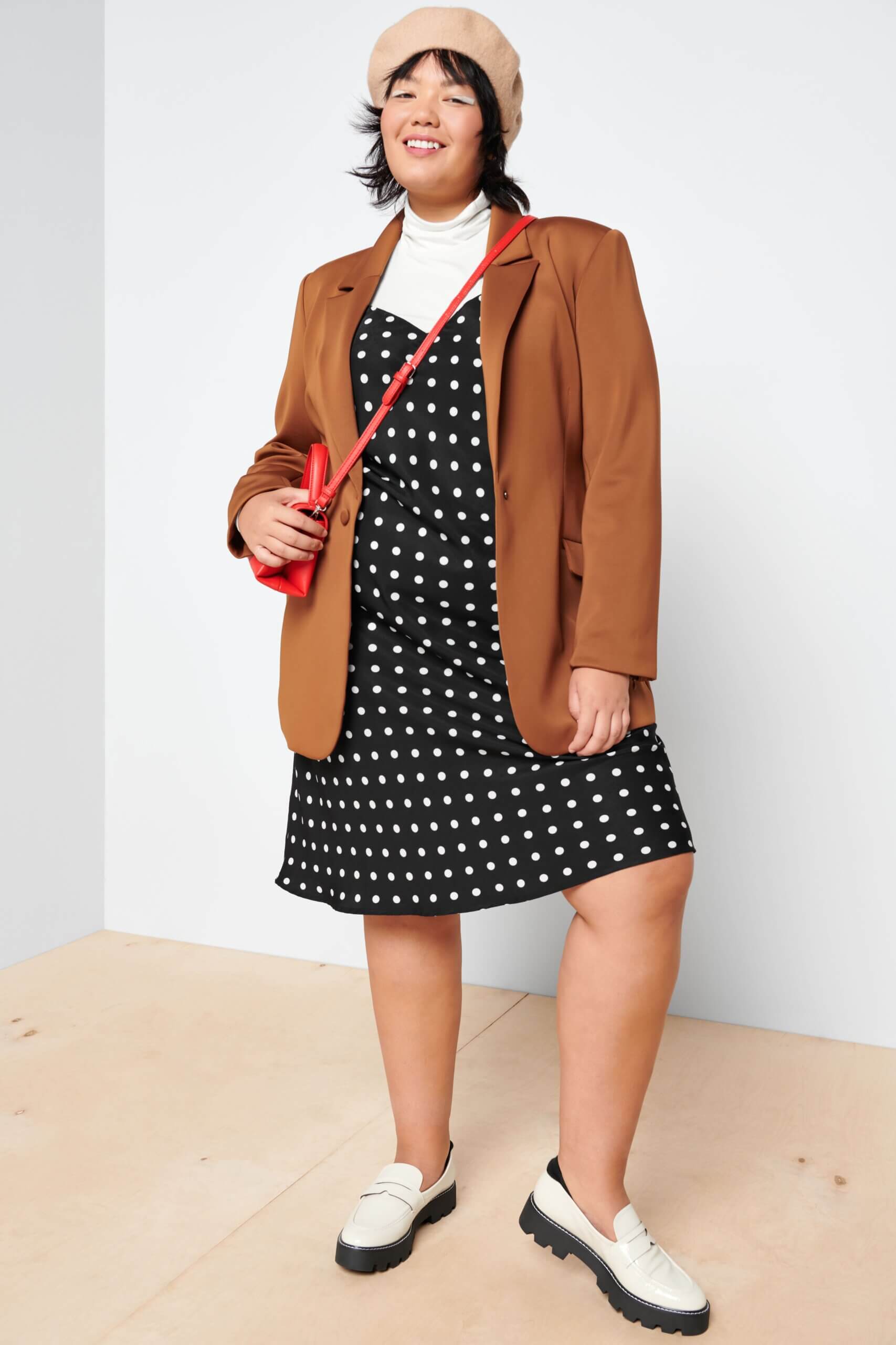 Stitch Fix model wearing women’s brown, black and white outfit with blazer, hat, polka dot dress, red bag and white loafers.