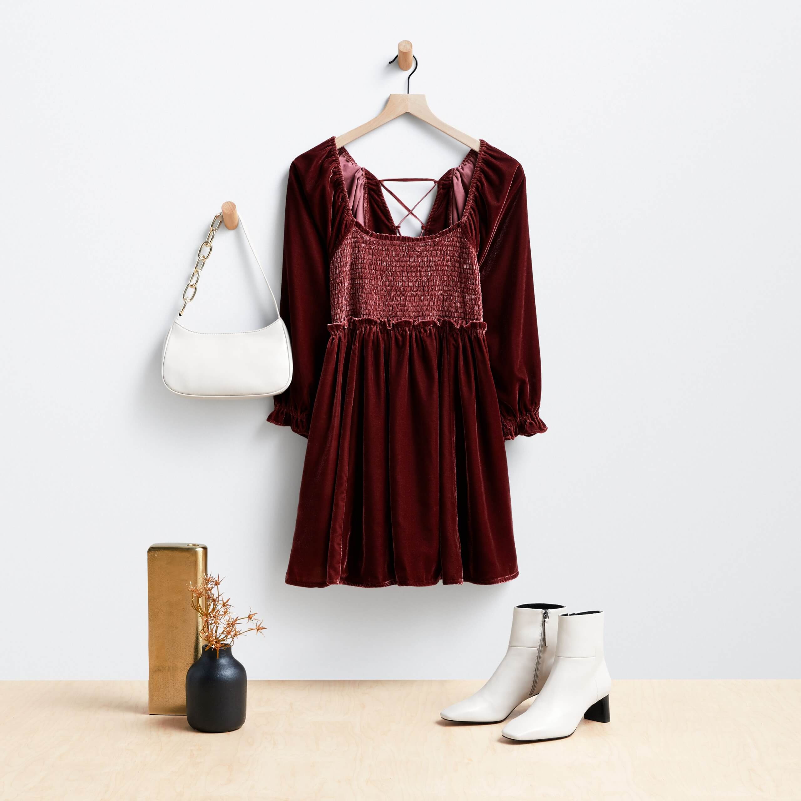 Stitch Fix women's burgundy velvet dress and white purse hanging on wooden hangers, next to white leather booties. 