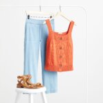 Stitch Fix Women’s cropped wide-leg jeans with orange button front tank top and brown sandals on a white stool.