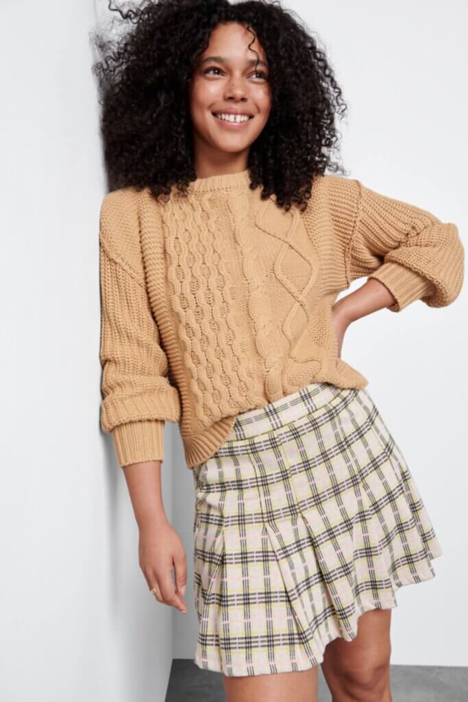 Stitch Fix Women’s model wearing a collegiate fashion outfit with a tan cable knit sweater and plaid skirt.