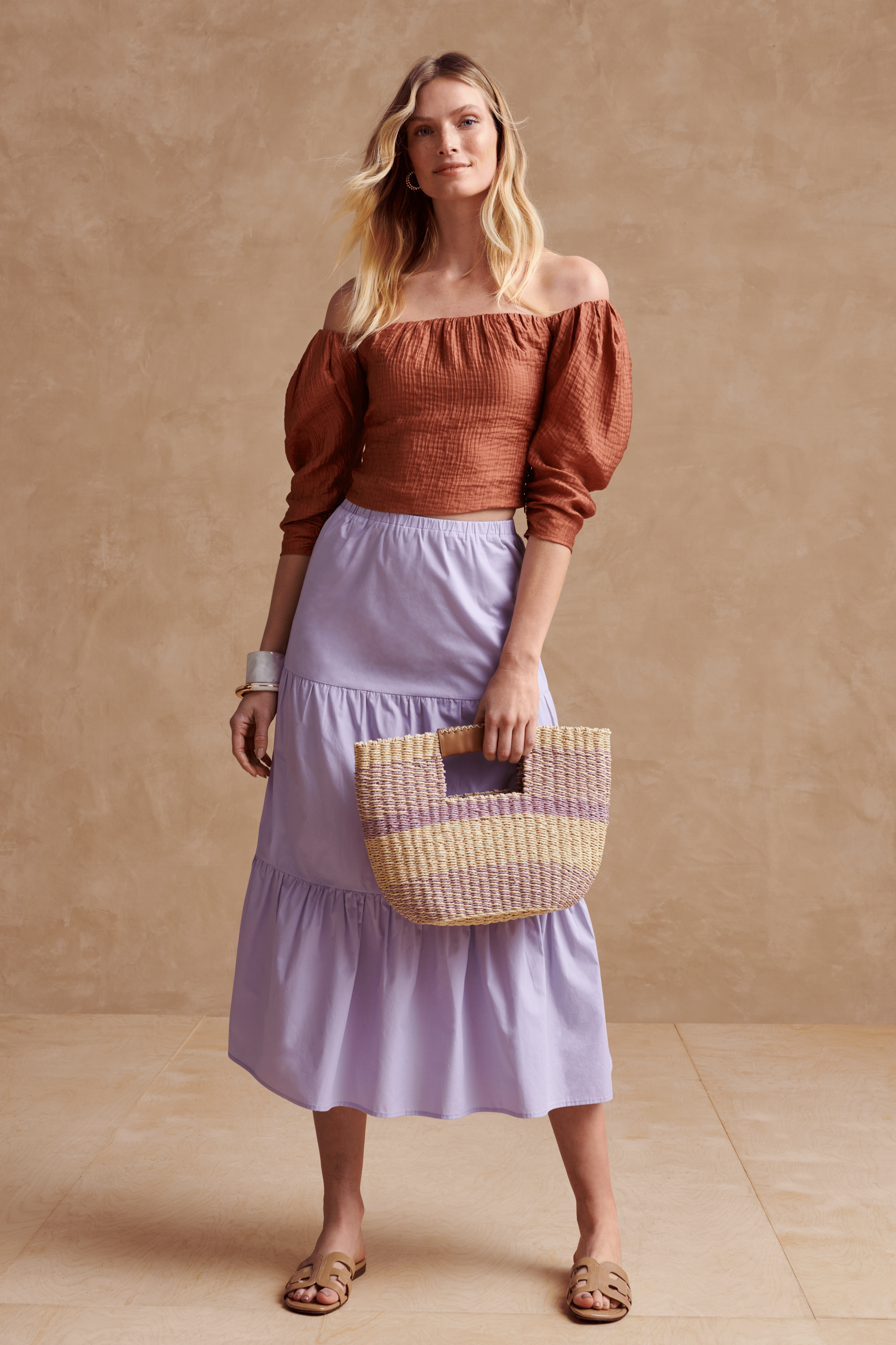 Stitch Fix model wearing women’s cottagecore aesthetic outfit with brown off-the-shoulder blouse, purple tiered midi skirt, rattan bag and tan sandals.
