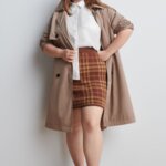 Stitch Fix Women’s model wearing a white button-up, brown plaid skirt, tan trench coat and black loafers.