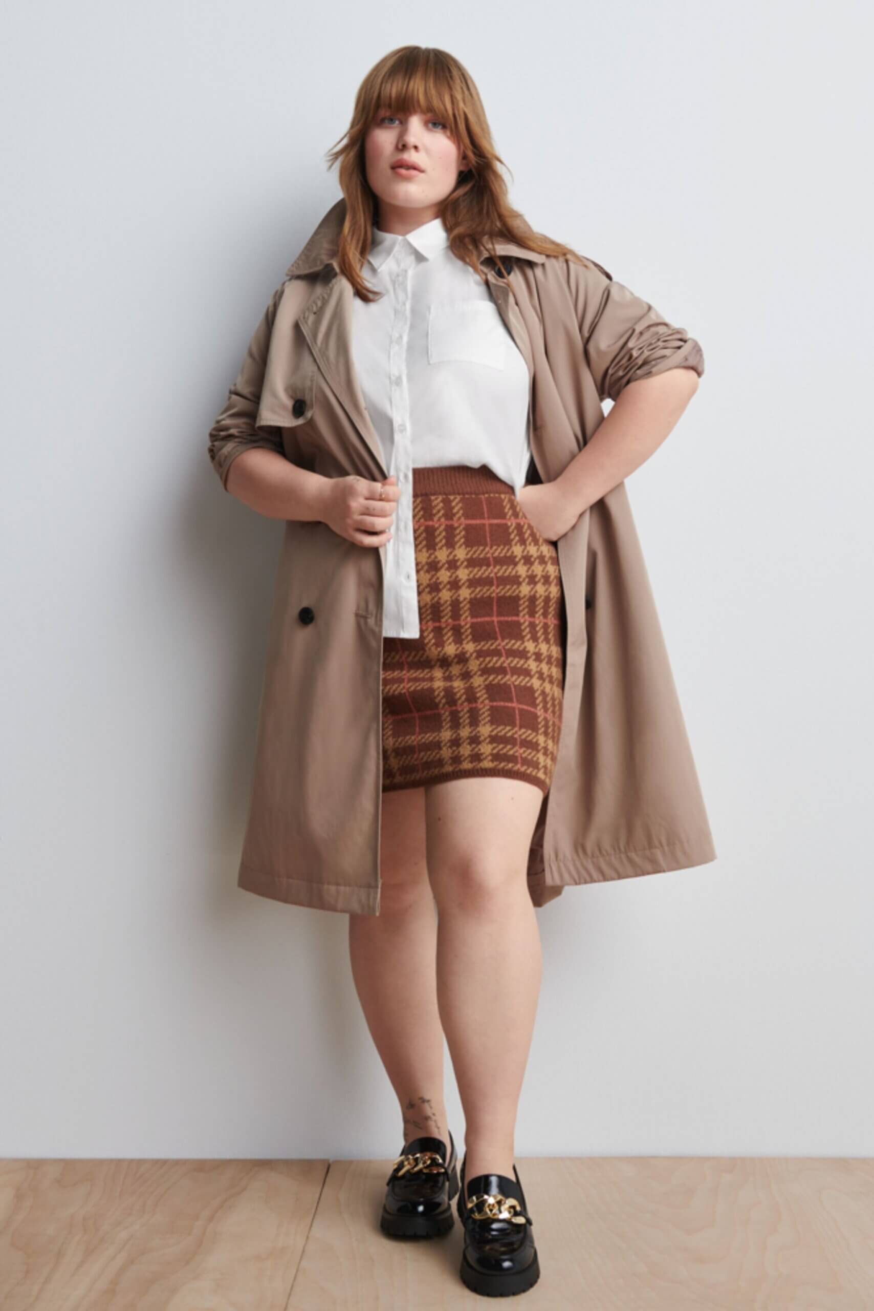 Stitch Fix Women’s model wearing a white button-up, brown plaid skirt, tan trench coat and black loafers.