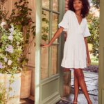 Stitch Fix model wearing women’s little white dress outfit with white dress, hoop earrings and heels by green door and flowers.