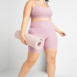Stitch Fix women's model wearing yoga outfit featuring pink sports bra, pink biker shorts, and grey sneakers holding a yoga mat.