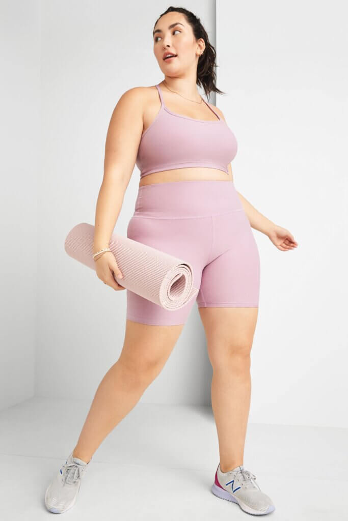 Stitch Fix women's model wearing yoga outfit featuring pink sports bra, pink biker shorts, and grey sneakers holding a yoga mat.
