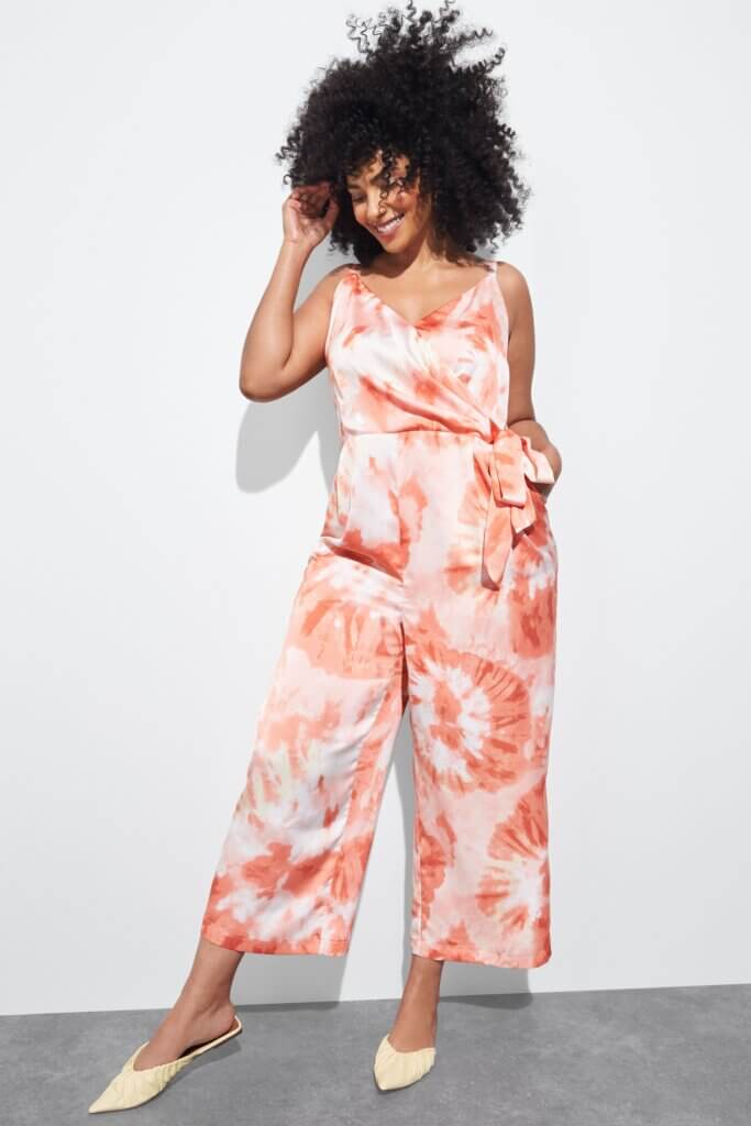 Stitch Fix model wearing women's semi-formal wedding attire featuring coral printed sleeveless jumpsuit and beige flats.