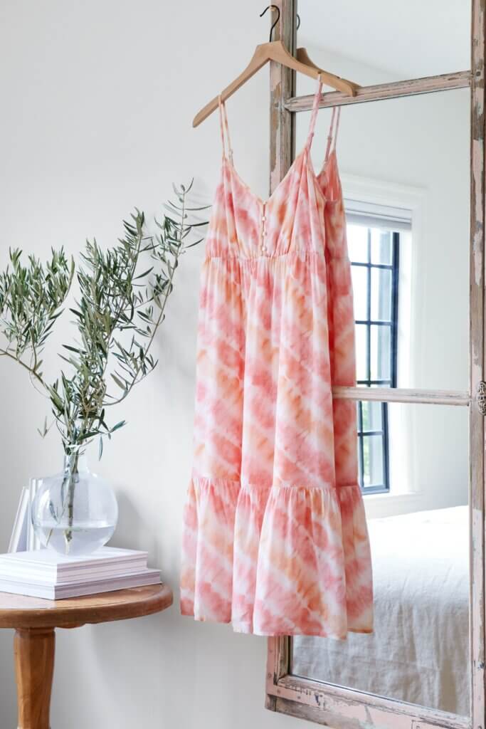 Stitch Fix women’s casual wedding beach attire with tie-dye dress hanging on mirror next to plant in vase and books on table.