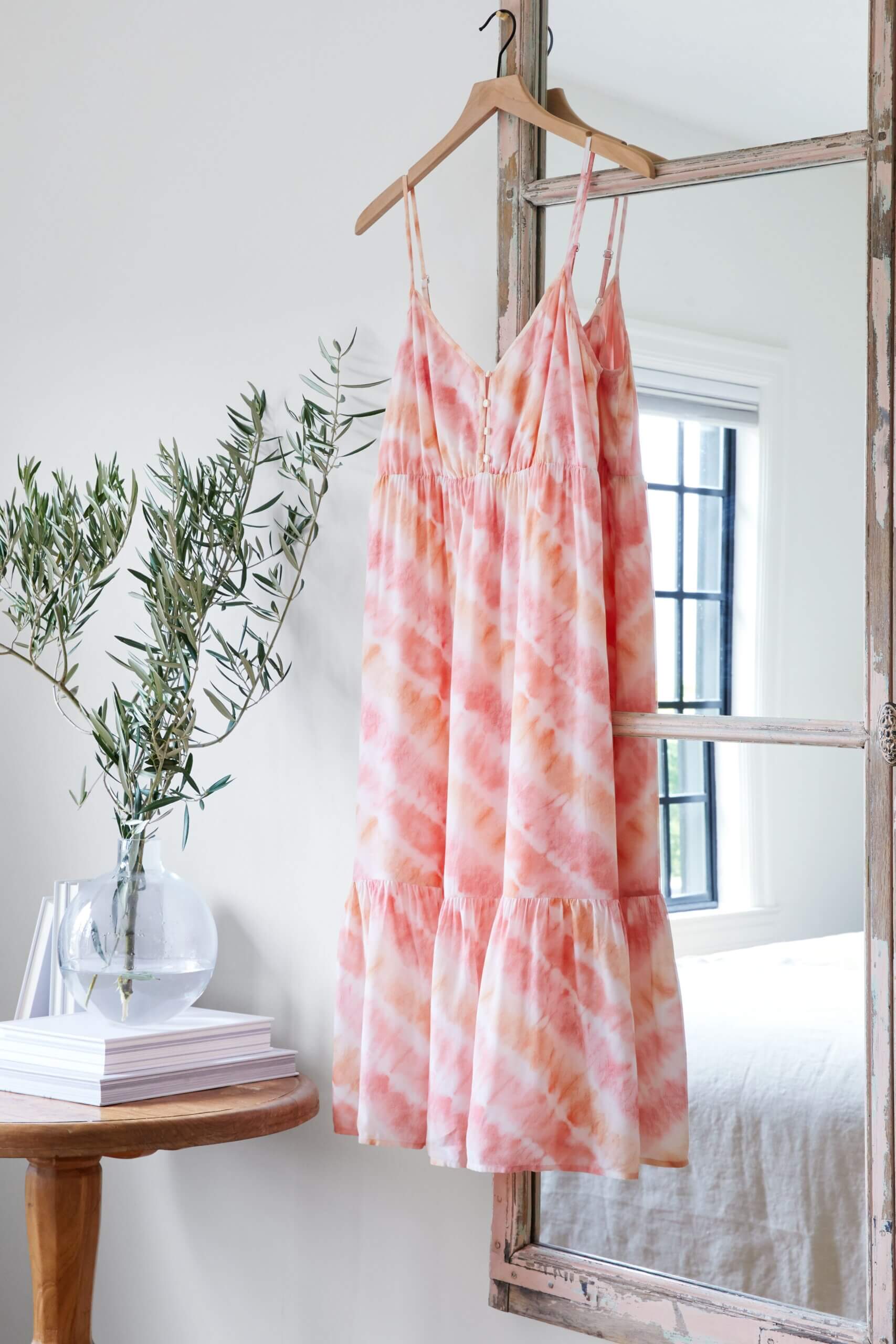 Stitch Fix women’s casual wedding beach attire with tie-dye dress hanging on mirror next to plant in vase and books on table.