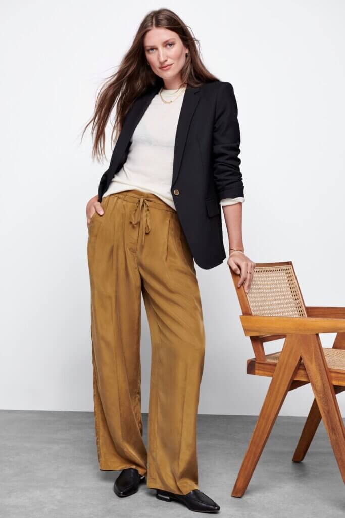 Stitch Fix model wearing women's 90s fashion outfit with white shirt, navy blazer, gold jewelry, satin pants and loafers leaning on chair.