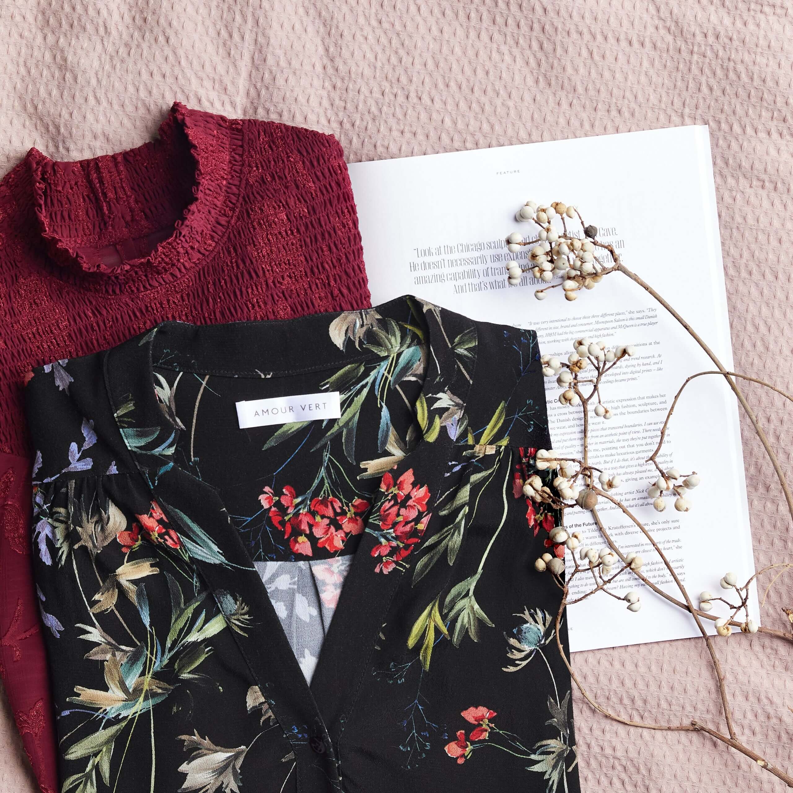 Stitch Fix women’s burgundy smocked top and black floral blouse laydown on white printed paper and blossoming twigs.