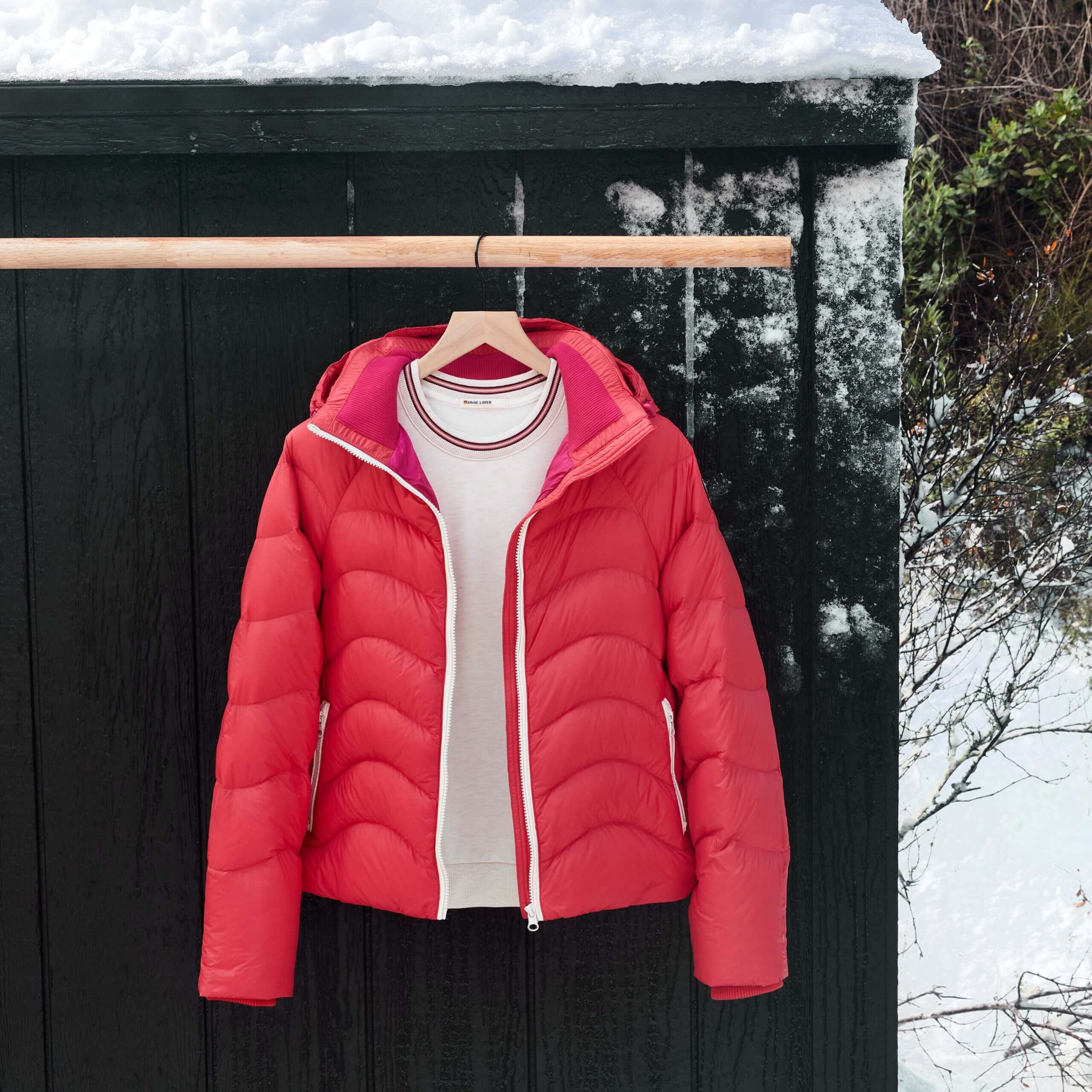 Stitch Fix women's red puffer jacket over white sweatshirt hanging on wooden pole outside of snowy cabin. 