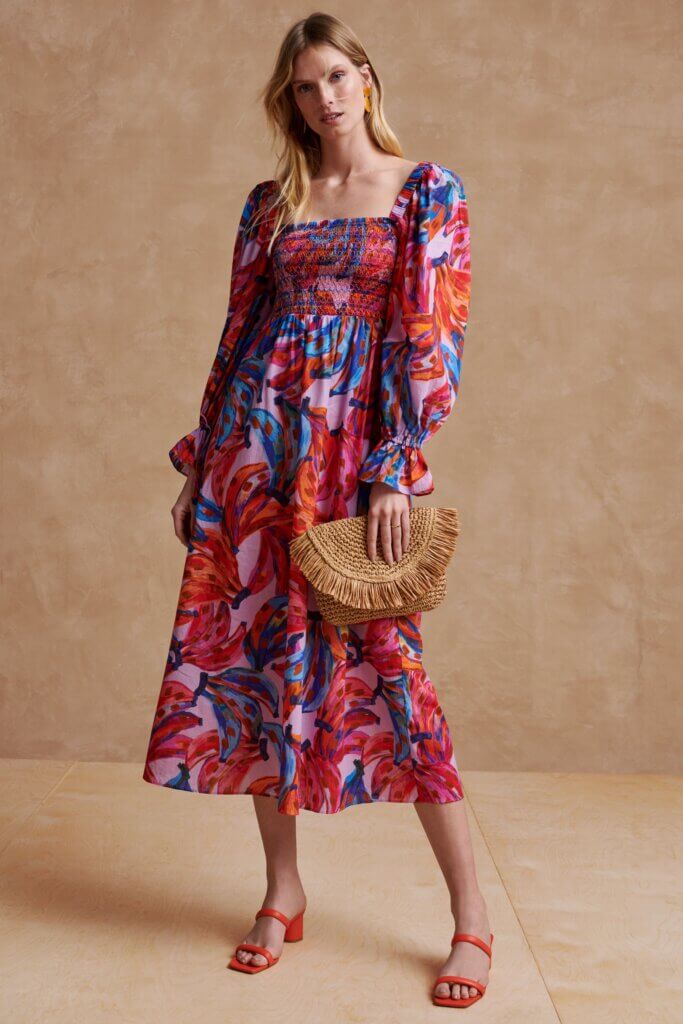Stitch Fix model wearing women’s beach wedding guest attire with multicolor print midi dress, rattan clutch and orange heels.