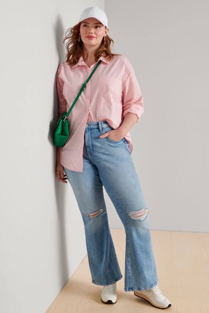 Stitch Fix model wearing women's 90s normcore fashion outfit with pink shirt, bootcut jeans, green bag and white sneakers and hat.