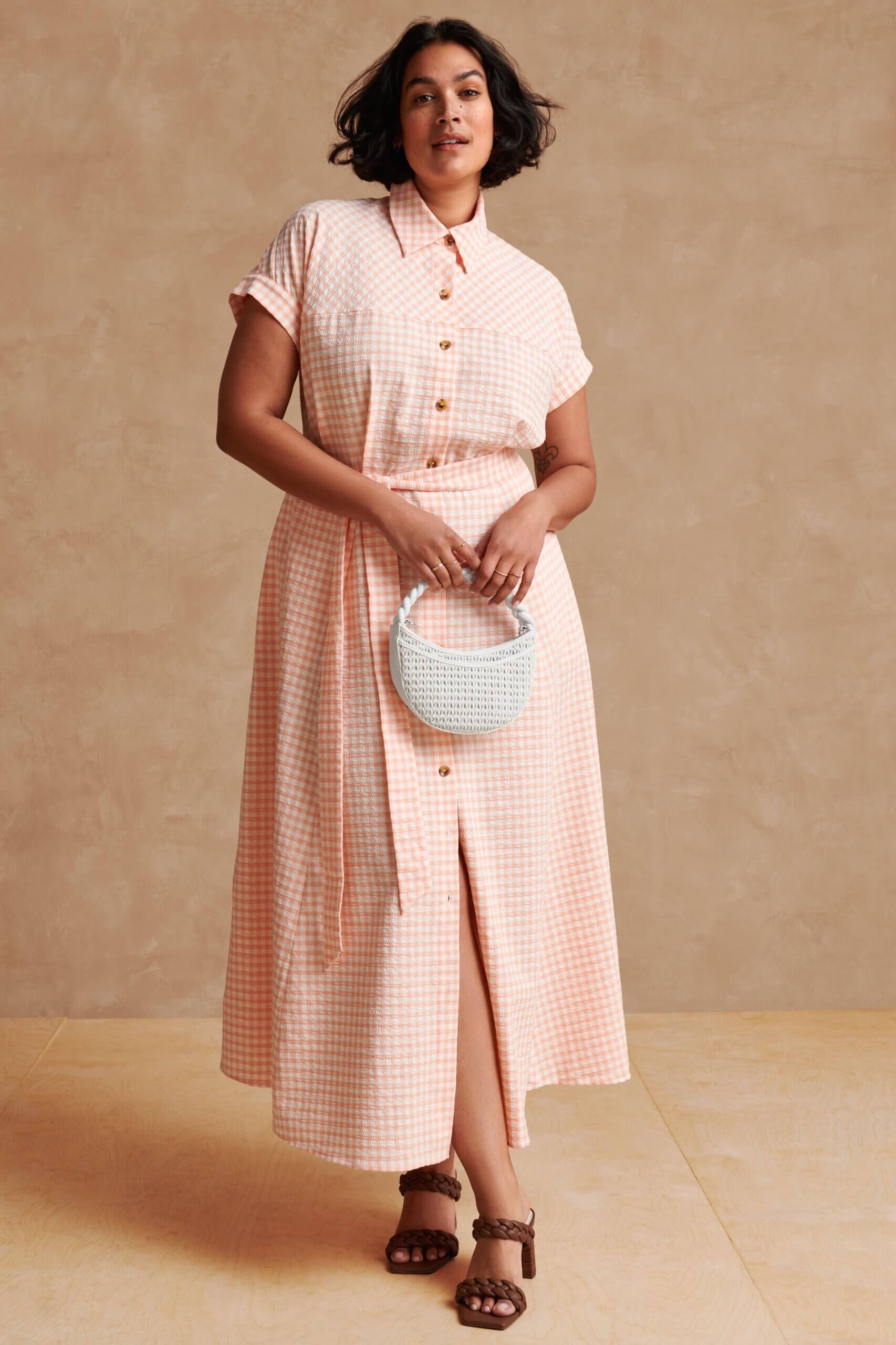 Stitch Fix model wearing women’s beach wedding guest attire with gingham dress, white handbag and brown braided heels.