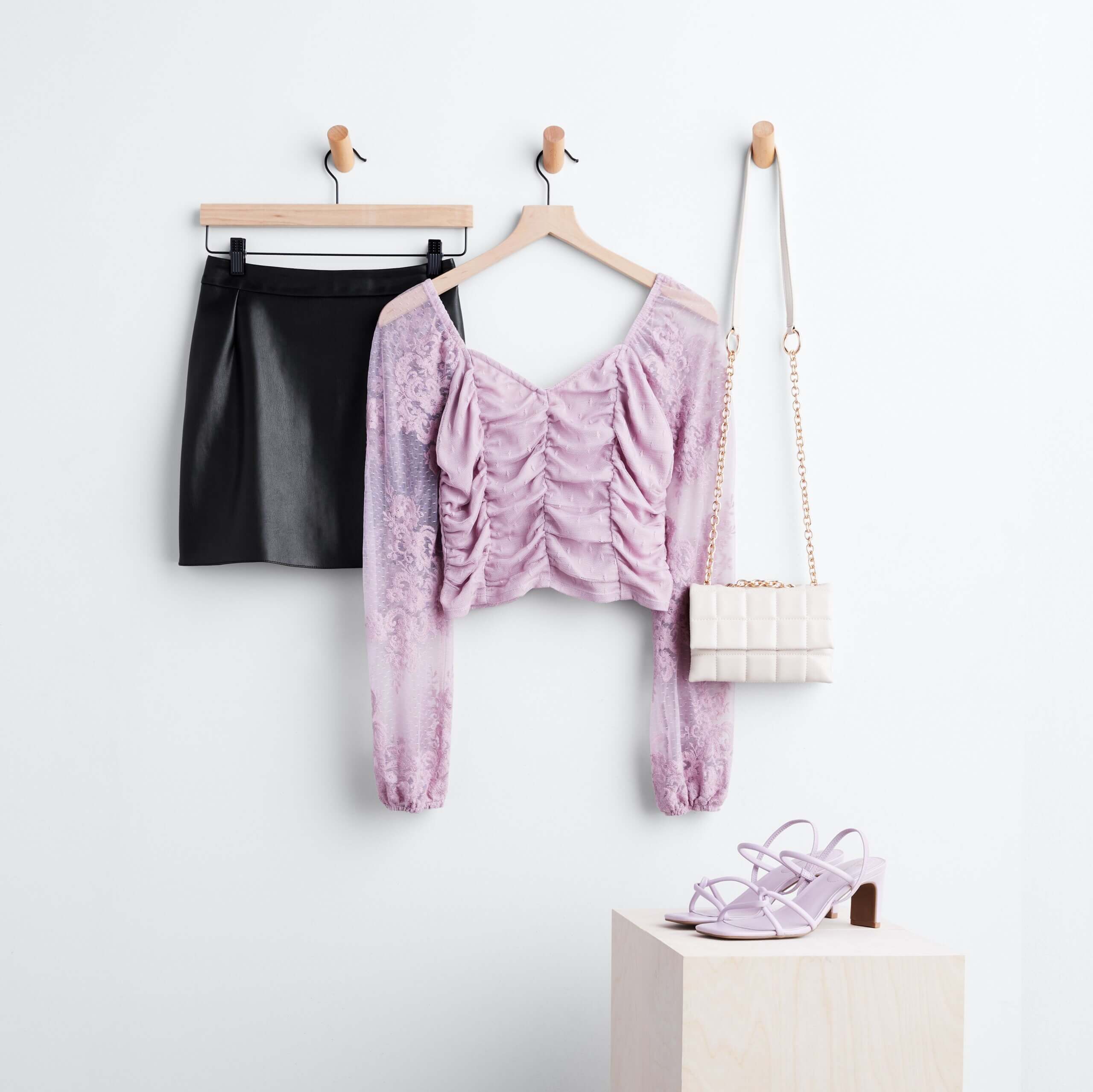 Stitch Fix women’s skirt outfit featuring black vegan leather skirt with tie waist, ruched lace long sleeve blouse, white crossbody purse on hangers and lavender heels on pedestal.