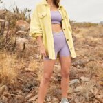 Stitch Fix model wearing women’s comfy road trip outfit with lilac and yellow bra and biker shorts, shacket, bucket hat and sneakers.