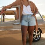 Stitch Fix model wearing women’s comfy road trip outfit with white cami, denim shorts, brown blazer and sandals standing next to car.