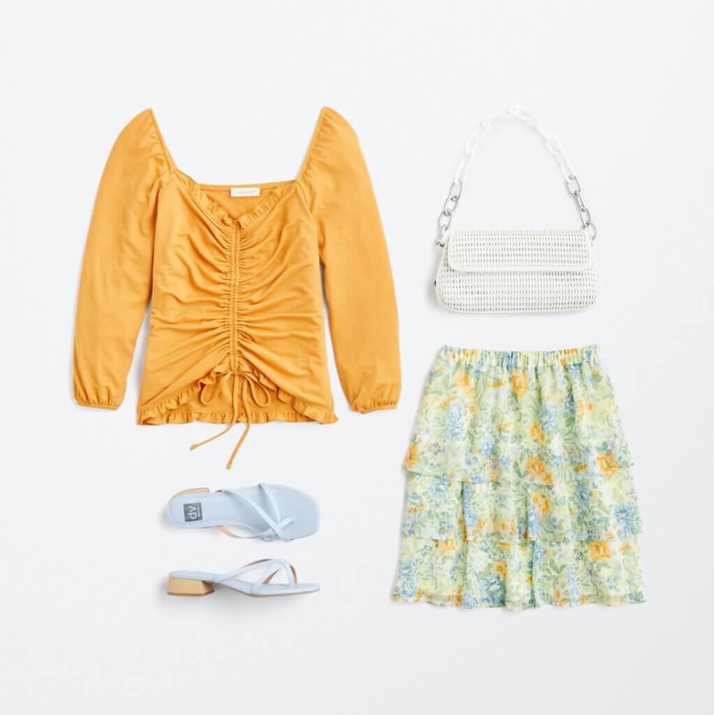 Stitch Fix women’s regencycore outfit featuring floral mini skirt, yellow ruched long-sleeve top, white purse and blue sandals.