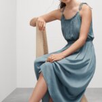 Stitch Fix model wearing women’s beach chic wedding attire with blue dress, gold accessories and sandals sitting on chair.