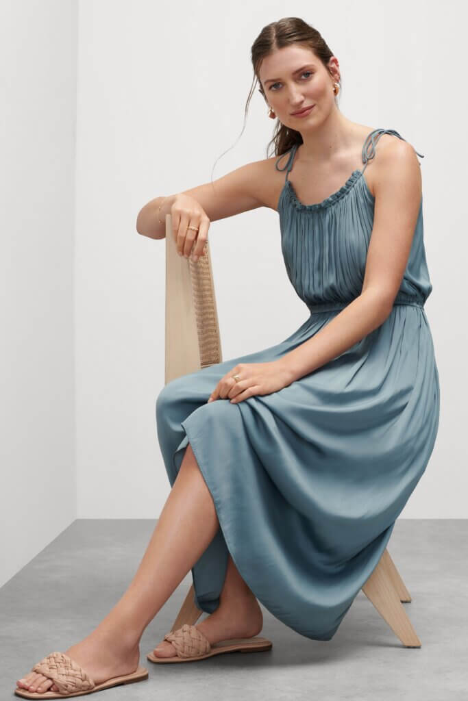 Stitch Fix model wearing women’s beach chic wedding attire with blue dress, gold accessories and sandals sitting on chair.