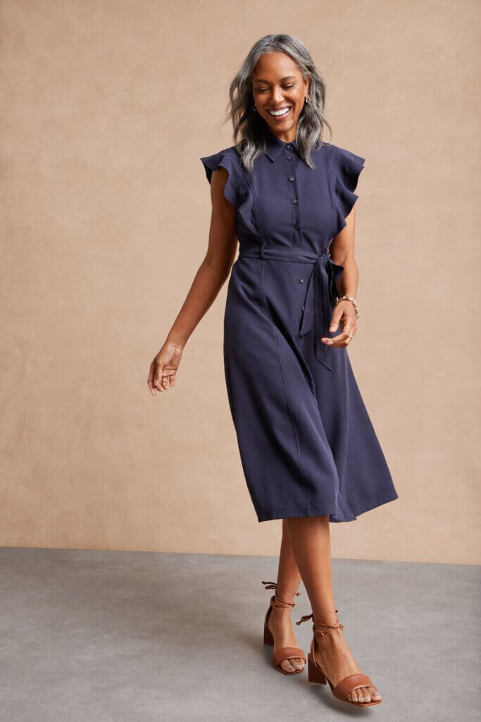 Stitch Fix women’s model wearing button-up accessorized navy blue dress for a wedding with brown sandals and gold bracelet.