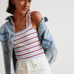 Stitch Fix model in women’s Americana aesthetic outfit featuring red, white and blue striped ruffle sleeve tank, denim jacket, white denim shorts and white baseball cap.