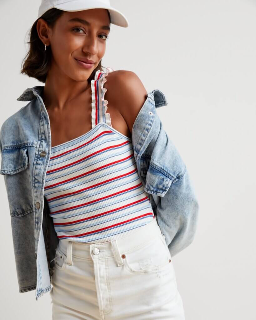 Stitch Fix model in women’s Americana aesthetic outfit featuring red, white and blue striped ruffle sleeve tank, denim jacket, white denim shorts and white baseball cap.
