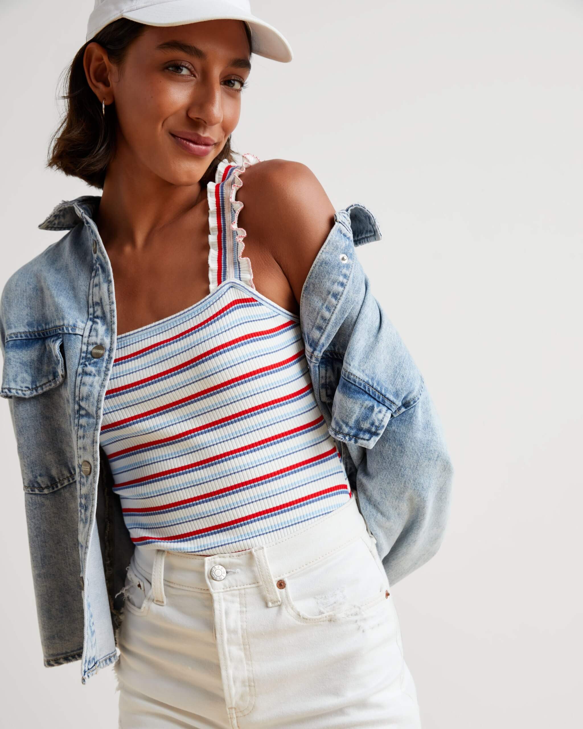 Stitch Fix model in women’s Americana aesthetic outfit featuring red, white and blue striped ruffle sleeve tank, denim jacket, white denim shorts and white baseball cap.