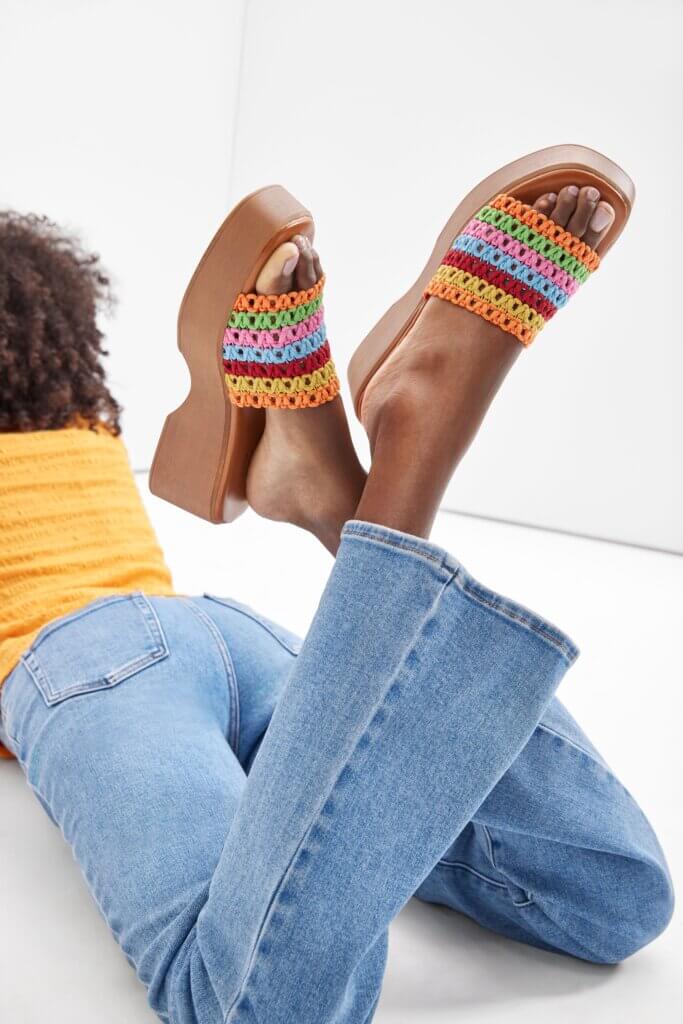 Back view of a woman’s legs in light wash denim jeans and colorful slip-on sandals.