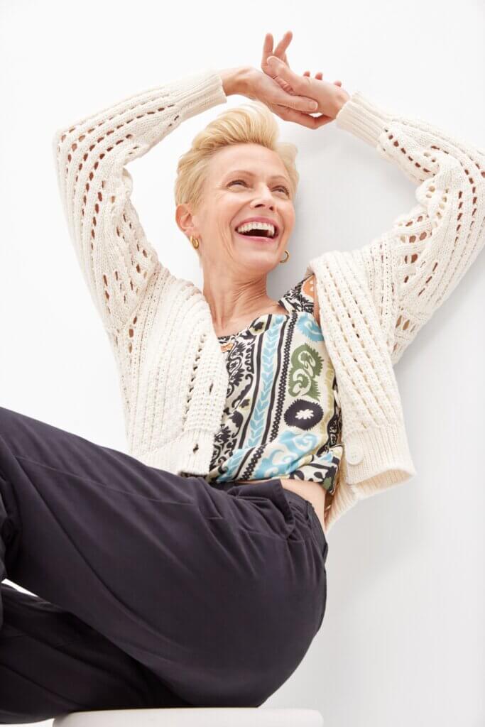 A woman sitting and smiling wearing a light sweater, colorful tank top and black pants.