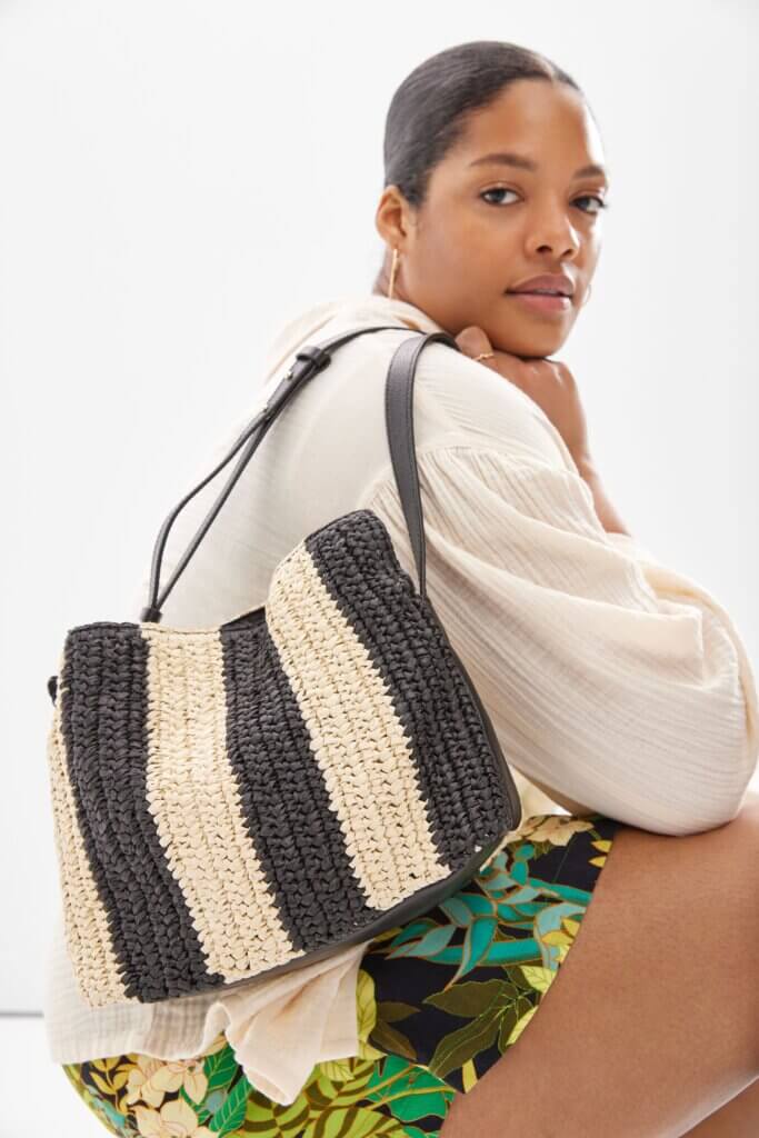 A woman sitting and looking into the camera wearing a natural feminine top, colorful shorts and holding a woven handbag.