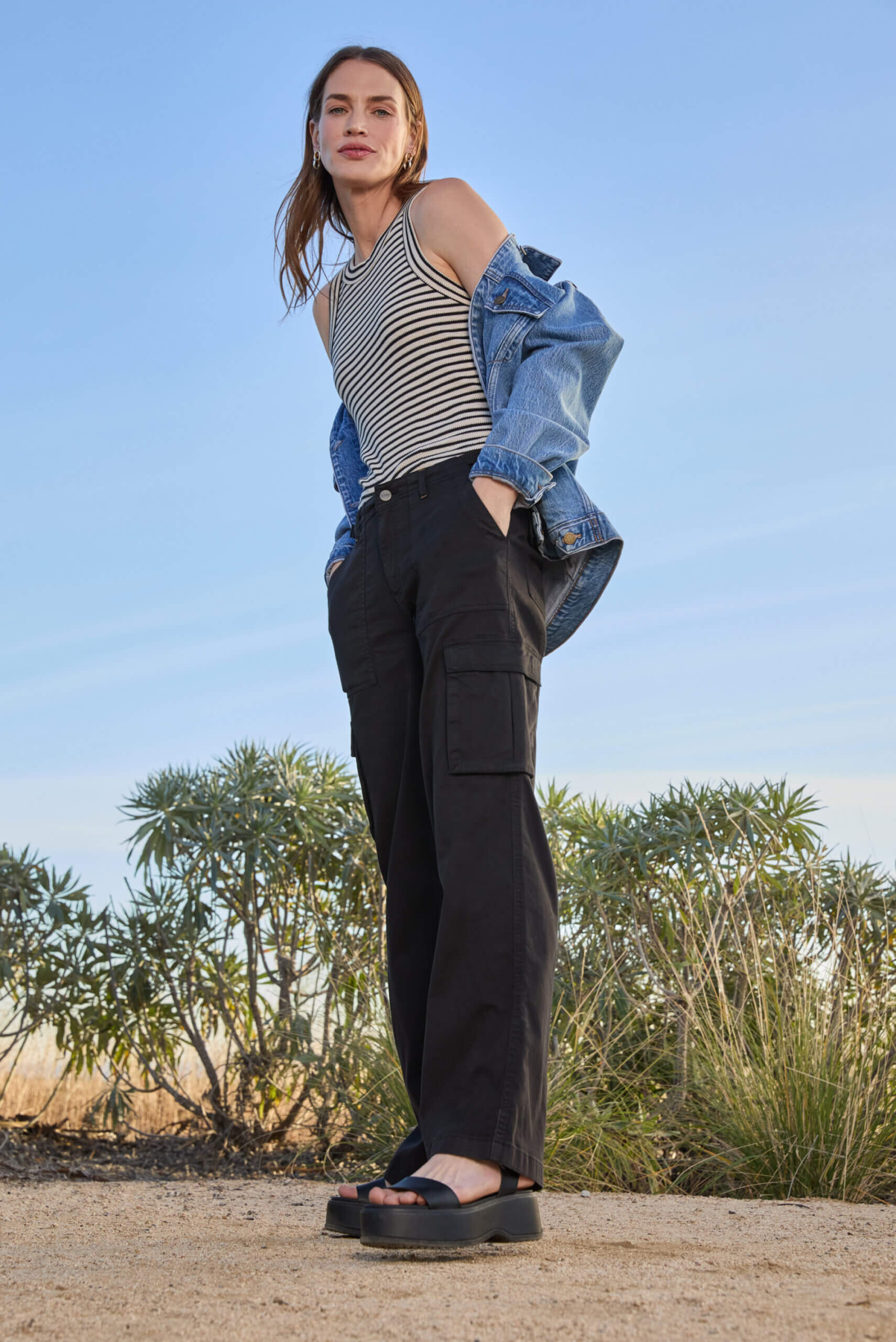 A woman in a black and white striped tank top, black cargo pants, a denim jacket, and black leather chunky sandals.